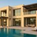 A modern ultra-luxury villa in Dubai featuring a minimalist architectural design with large glass windows, an infinity pool, sun loungers, and landscaped palm surroundings overlooking a tranquil lagoon at sunset.
