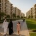 A family walks along landscaped pathways lined with modern low-rise apartment buildings in Dubai South, showcasing a vibrant residential community with green spaces and contemporary architecture under warm afternoon sunlight.